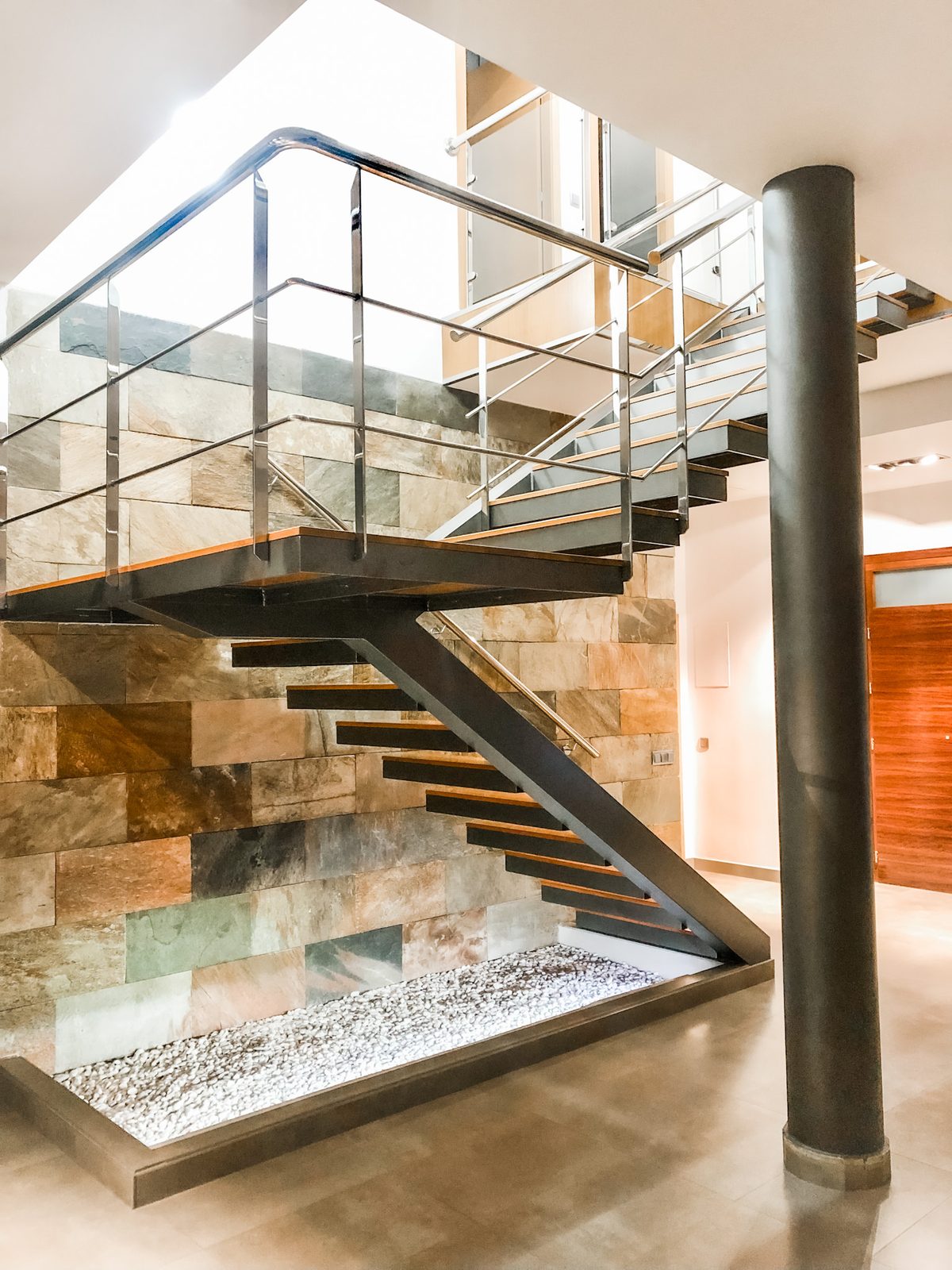 The open-tread steel and timber staircase against the split-face stone wall, seen from below with the mezzanine above