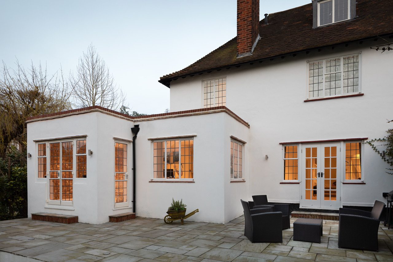 Rear exterior at dusk showing the rendered extension, leaded windows, and garden terrace illuminated from within