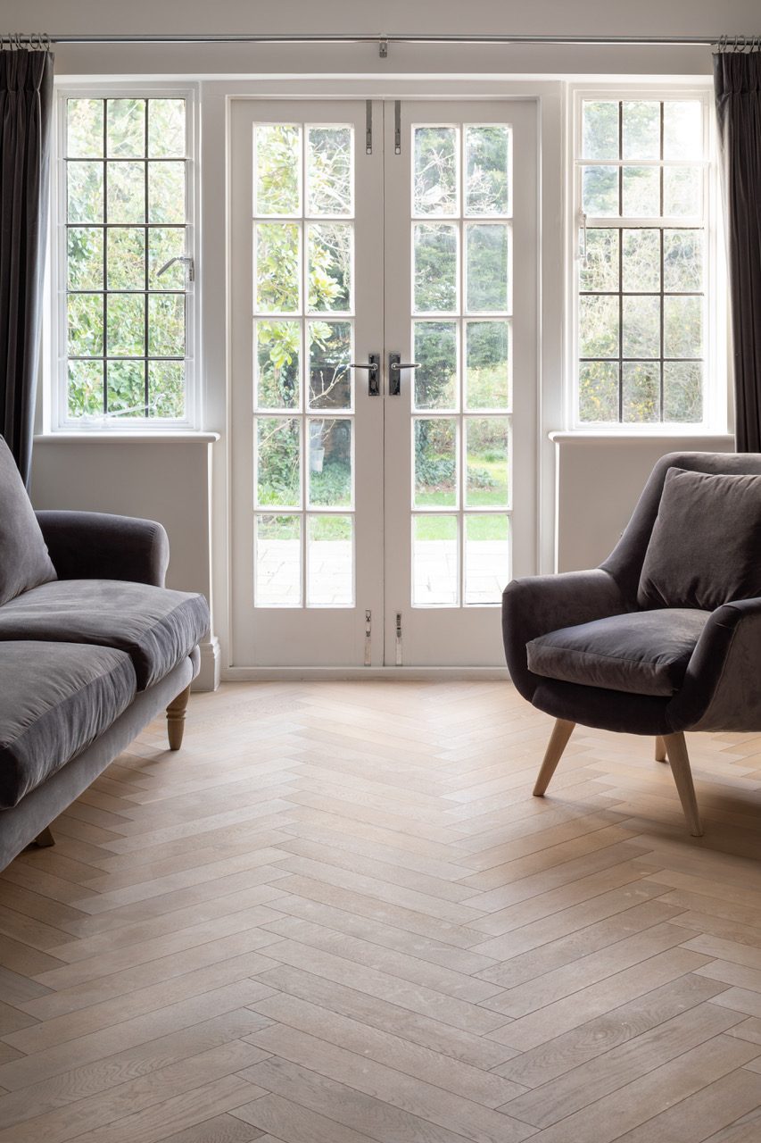 Living room with herringbone oak flooring, grey velvet sofa and armchair, facing leaded French doors onto the garden